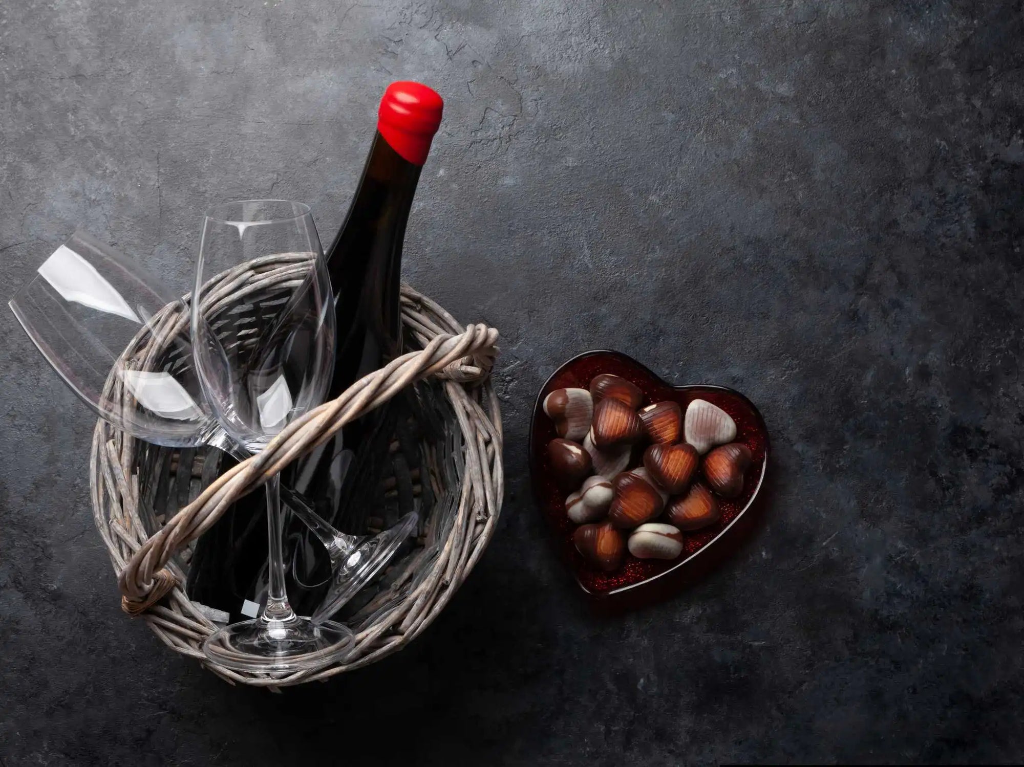 Korb mit Weinflasche alkoholfrei und zwei Gläsern, daneben herzförmiger Teller mit Pralinen auf dunklem Hintergrund.