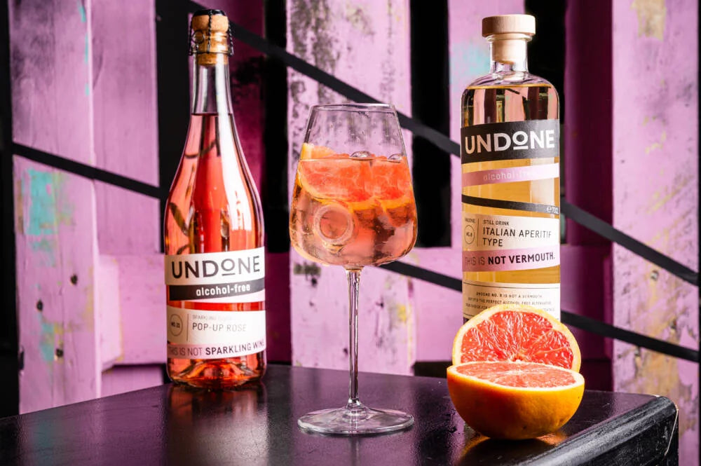 Undone alkoholfreier Pop-Up Rosé und Italian Aperitif Type auf einem Tisch mit einem Glas Getränk und einer halben Grapefruit.