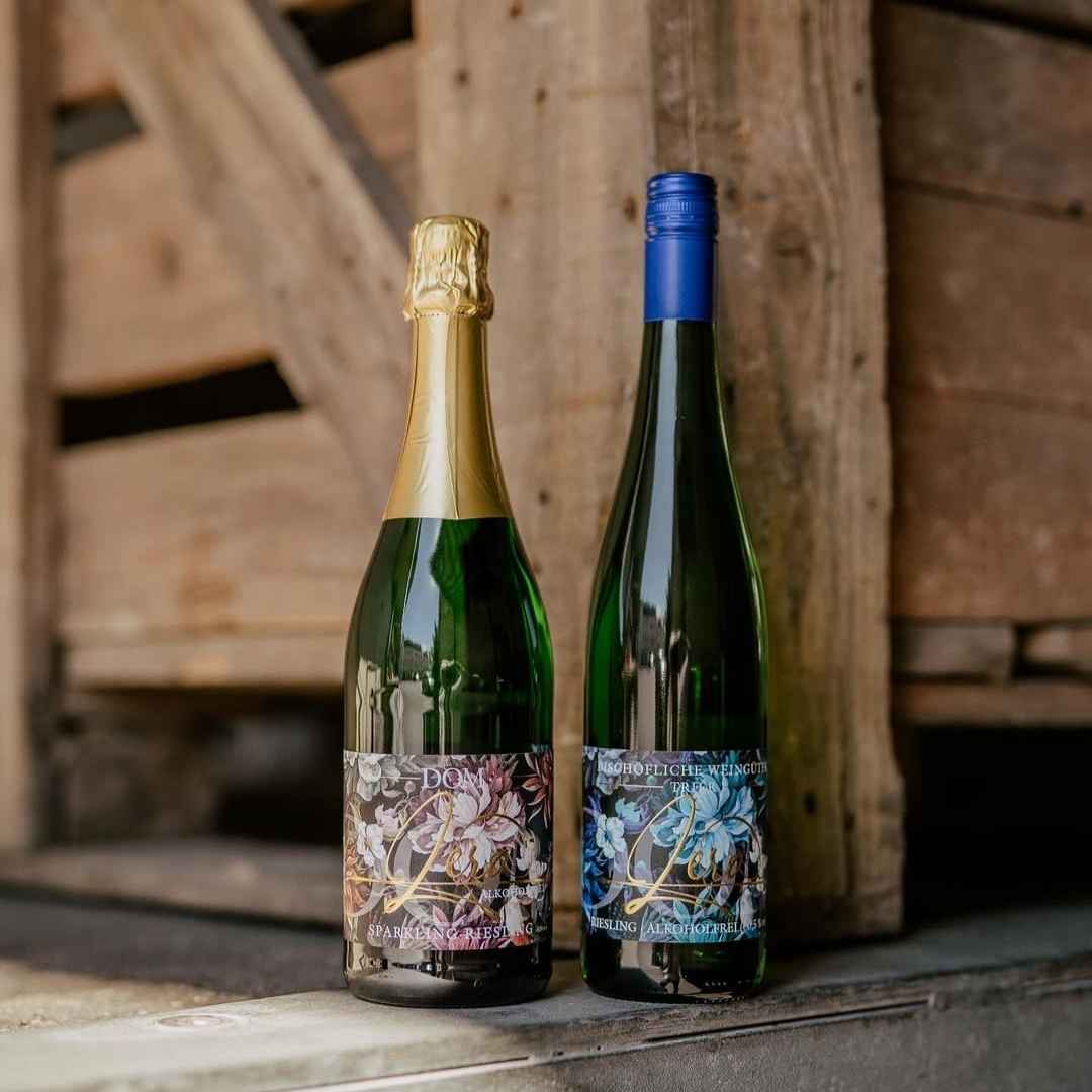 Zwei Flaschen DOM Zero alkoholfrei: Sparkling Riesling mit goldener Kapsel und stiller Riesling mit blauem Schraubverschluss vor rustikalem Holz-Hintergrund