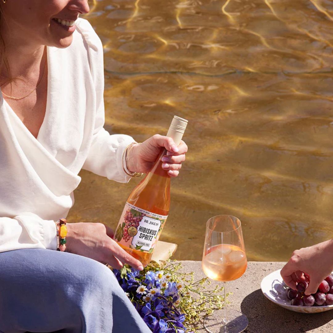 Frau hält Dr. Jaglas Hibiskus Spritz in der Hand beim sommerlichen Picknick am Wasser – mit Weinglas und roten Trauben