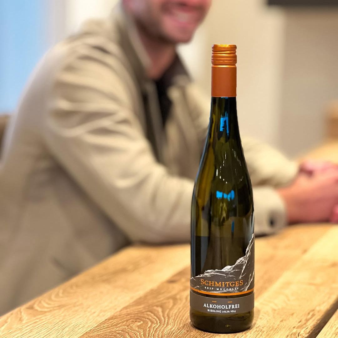 Flasche Schmitges Riesling alkoholfrei auf einem Holztisch, im Hintergrund sitzt unscharf ein lächelnder Mann in heller Jacke.