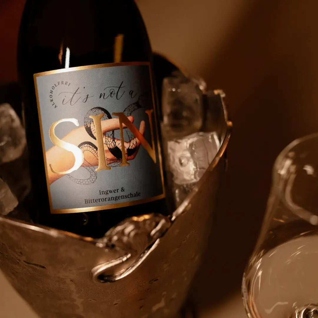 Alkoholfreie Sekt-Alternative „It’s not a SIN“ Ingwer & Bitterorangenschale im Sektkühler, stilvoll präsentiert mit Gläsern auf gedecktem Tisch